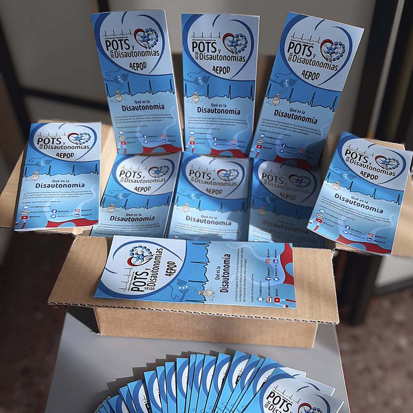 Qué hacemos Foto de una caja de cartón de un palmo de altura llena de trípticos azules de AEPOD titulado “Qué es la Disautonomía”. Unos están dentro de la caja y otros de pie apoyados en las solapas abiertas. Está encima de una mesita auxiliar y le dan los rayos de sol de la mañana de la ventana.