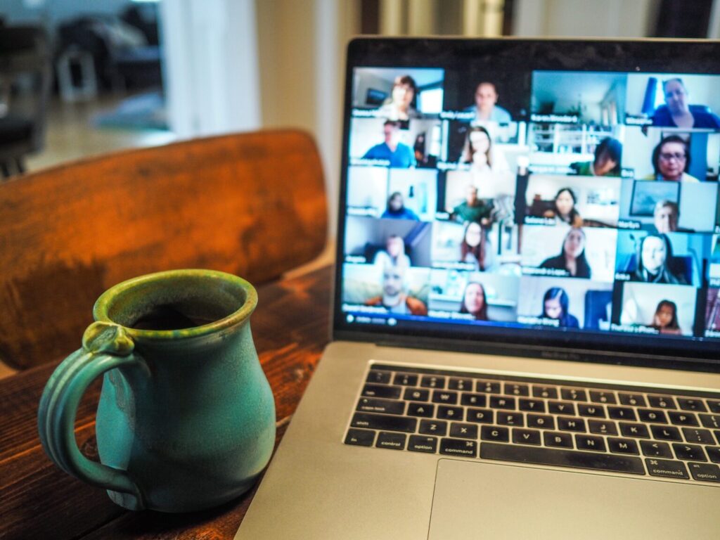 Foto de un ordenador portátil abierto encima de una mesa mostrando en la pantalla una videoconferencia con 25 recuadros de participantes borrosos. Al lado, una taza con asa color verde.
