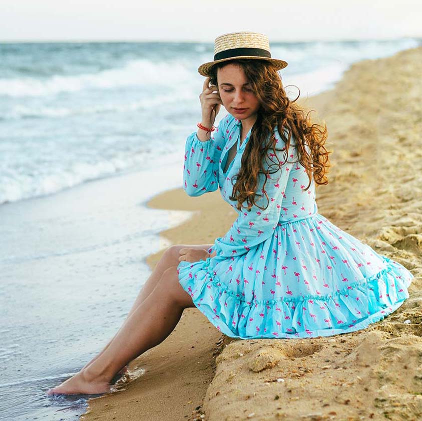 Foto de una mujer joven con pelo largo rizado castaño, sombrero de paja y vestido azul hasta las rodillas, sentada en un montículo de arena de playa con los pies dentro del agua en la orilla del mar. De fondo a lo lejos el horizonte.